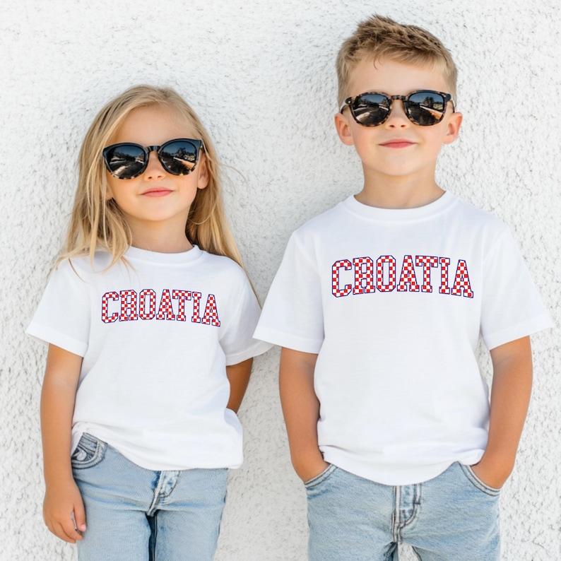 May include: Two children wearing white t-shirts with the word "CROATIA" in red and white checkered letters. Both children are wearing sunglasses and blue jeans. The background is a plain white wall.