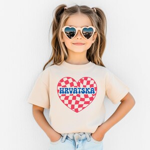 Puede incluir: Una ni&ntilde;a peque&ntilde;a con una camiseta beige claro con un dise&ntilde;o de coraz&oacute;n a cuadros rojos y blancos y la palabra "HRVATSKA" en letras azules. Lleva gafas de sol en forma de coraz&oacute;n y tiene el pelo recogido en dos coletas.