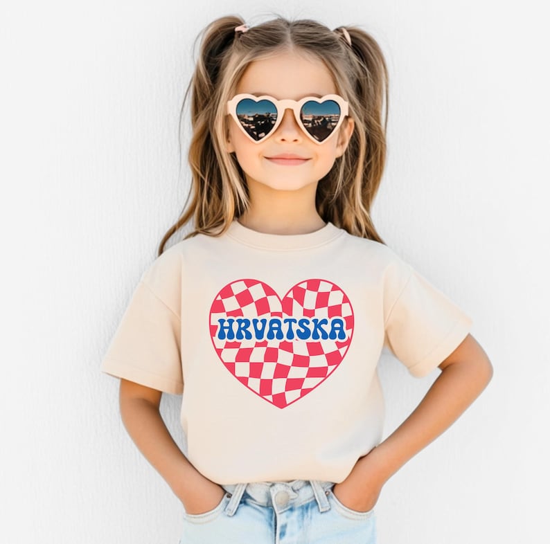 Puede incluir: Una ni&ntilde;a peque&ntilde;a con una camiseta beige claro con un dise&ntilde;o de coraz&oacute;n a cuadros rojos y blancos y la palabra "HRVATSKA" en letras azules. Lleva gafas de sol en forma de coraz&oacute;n y tiene el pelo recogido en dos coletas.