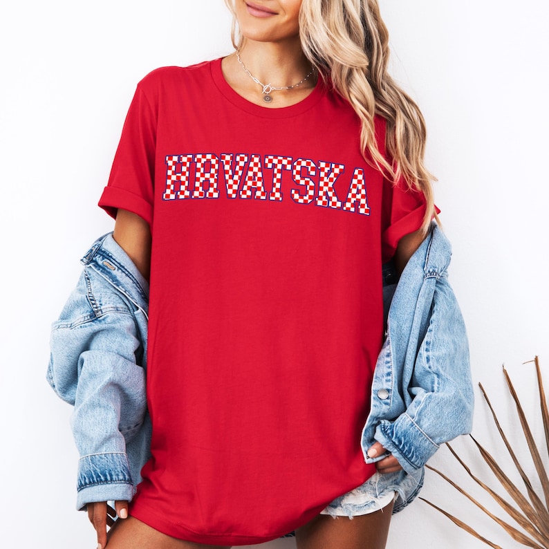Pode incluir: Uma camiseta vermelha com a palavra "HRVATSKA" em um padr&atilde;o xadrez vermelho, branco e azul. A camiseta tem mangas curtas e &eacute; usada com uma jaqueta jeans. O tema geral &eacute; patri&oacute;tico.