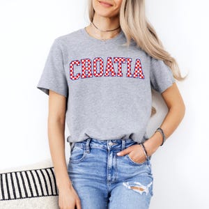 May include: A gray t-shirt with the word "CROATIA" in red and white checkered letters. The shirt is short-sleeved and has a crew neck. The person is wearing blue jeans.