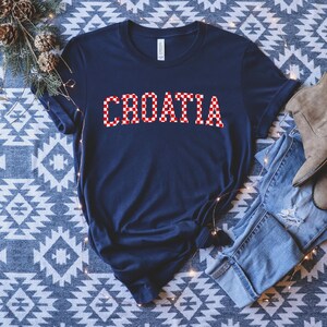May include: Navy blue t-shirt with the word "CROATIA" in a red and white checkered pattern. The shirt is made of a soft material and has short sleeves. The shirt is folded on a patterned surface.