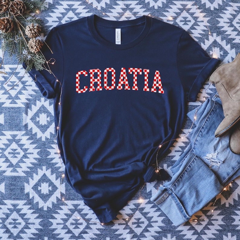 May include: Navy blue t-shirt with the word "CROATIA" in a red and white checkered pattern. The shirt is made of a soft material and has short sleeves. The shirt is folded on a patterned surface.