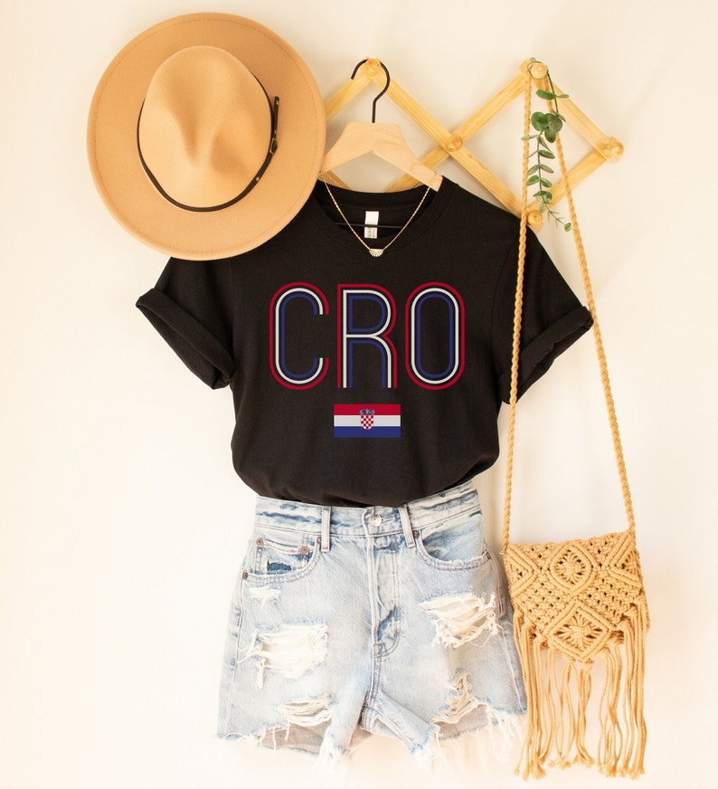 May include: Black t-shirt with the word "CRO" in red, white, and blue lettering and the Croatian flag below. The shirt is hanging on a wooden hanger with a straw hat and a woven bag.