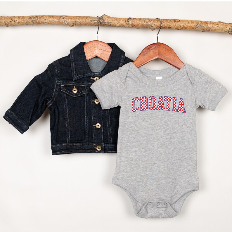 May include: A dark blue denim jacket and a gray baby onesie. The onesie has the word "CROATIA" in red and white checkered letters. Both items are hanging on wooden hangers from a branch.