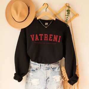 Pu&ograve; includere: Felpa nera con testo rosso che recita "VATRENI REPUBLIKA HRVATSKA". La felpa &egrave; appesa a un appendiabiti di legno ed &egrave; abbinata a un cappello marrone e una gonna di jeans.