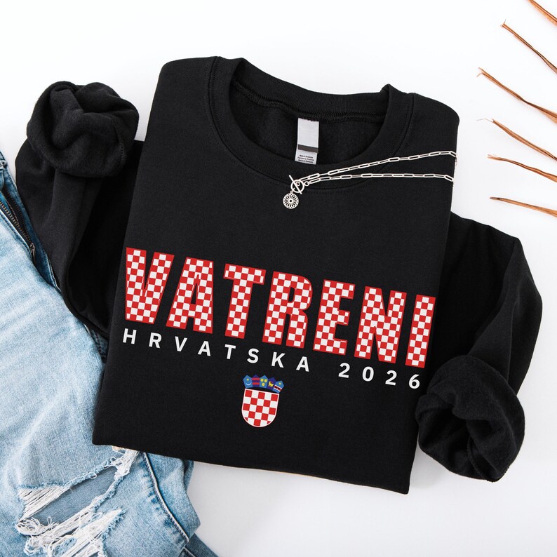 May include: Black sweatshirt featuring the word "VATRENI" in red and white checkered letters. Below the word is "HRVATSKA 2026" and a Croatian coat of arms. A silver necklace with a circular pendant is on the sweatshirt.