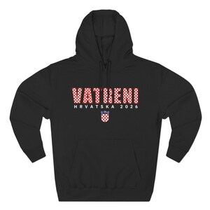May include: Black hooded sweatshirt with the word "VATRENI" in a red and white checkered pattern. Below the word is "HRVATSKA 2026" and a Croatian coat of arms. The sweatshirt has a drawstring hood and long sleeves.