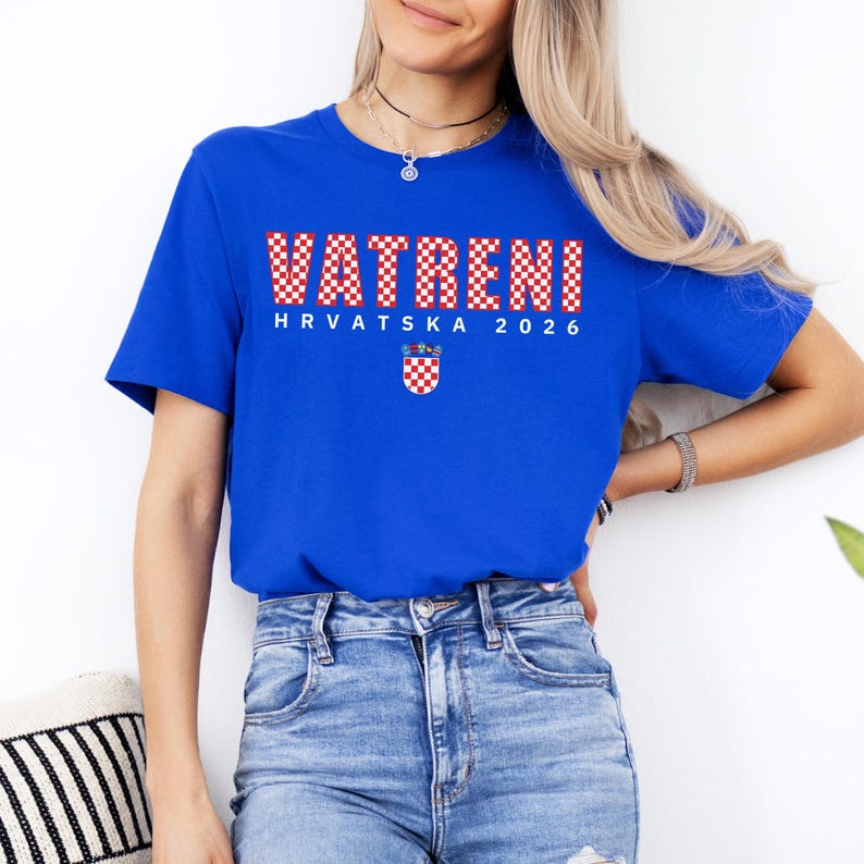 May include: Royal blue t-shirt with the word "VATRENI" in a red and white checkered pattern. Below the word is "HRVATSKA 2026" and a Croatian coat of arms. The shirt is made of a soft material.