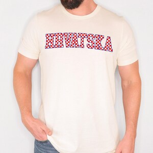 May include: Cream-coloured t-shirt with the word "HRVATSKA" in a red, white, and blue chequered pattern. The text is outlined in blue. The shirt has short sleeves and a crew neck.