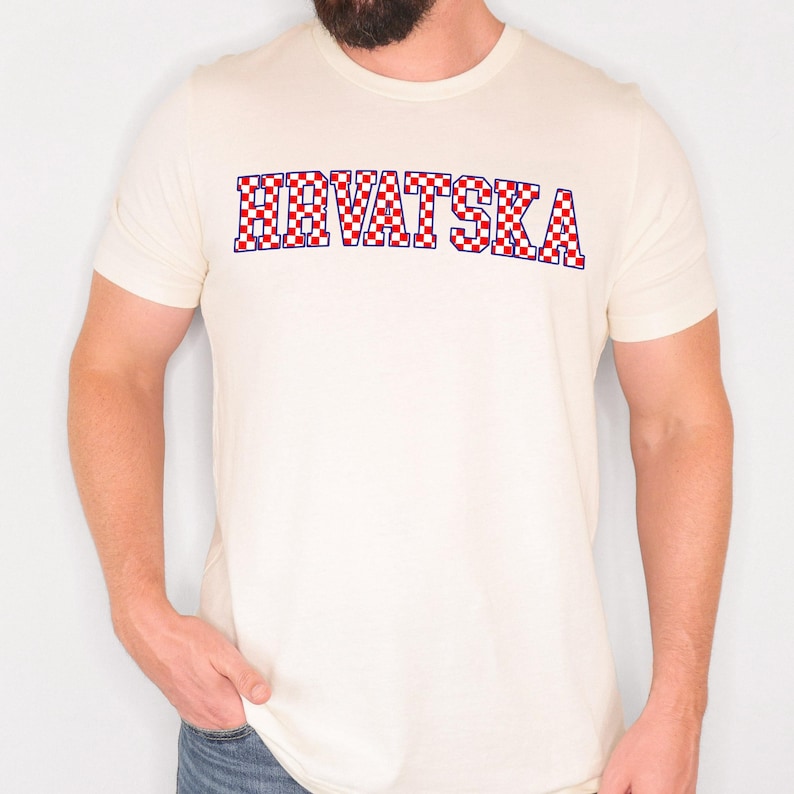 May include: Cream-coloured t-shirt with the word "HRVATSKA" in a red, white, and blue chequered pattern. The text is outlined in blue. The shirt has short sleeves and a crew neck.