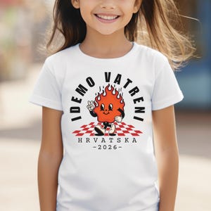 May include: White t-shirt featuring a cartoon flame character with the text "IDEMO VATRENI" in a semi-circle above the flame. The flame is on a red and white checkered pattern with the text "HRVATSKA - 2026-" below.