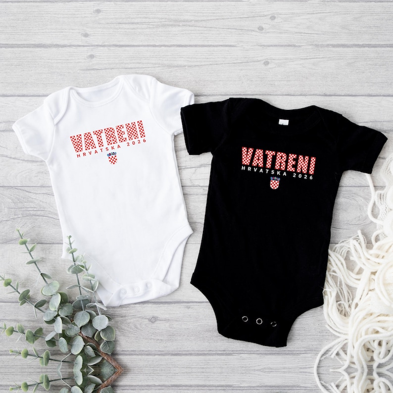 K&ouml;nnte beinhalten: Zwei Baby-Bodys, einer wei&szlig; und einer schwarz, werden auf einer hellen Holzoberfl&auml;che gezeigt. Jeder Body hat das Wort "VATRENI" in rot karierten Buchstaben, darunter "HRVATSKA 2026" und ein kroatisches Wappen.