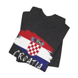 K&ouml;nnte beinhalten: Ein dunkelgraues T-Shirt mit einem kroatischen Flaggen-Design in Wei&szlig;, Rot und Blau. Die Flagge ist im Pinselstrich-Stil gemalt und das Wort "CROATIA" ist in Wei&szlig; unter der Flagge geschrieben.