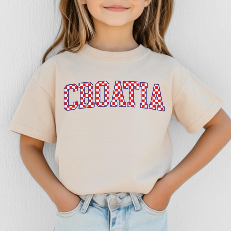 May include: A beige t-shirt with the word "CROATIA" in large, bold letters across the front. The letters are filled with a red and white checkered pattern, outlined in blue. The shirt is short-sleeved.