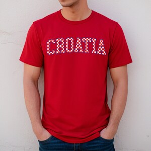 May include: A red t-shirt with the word "CROATIA" across the chest. The text is in a white, red, and blue checkered pattern, reminiscent of the Croatian flag. The shirt has short sleeves and a crew neck.