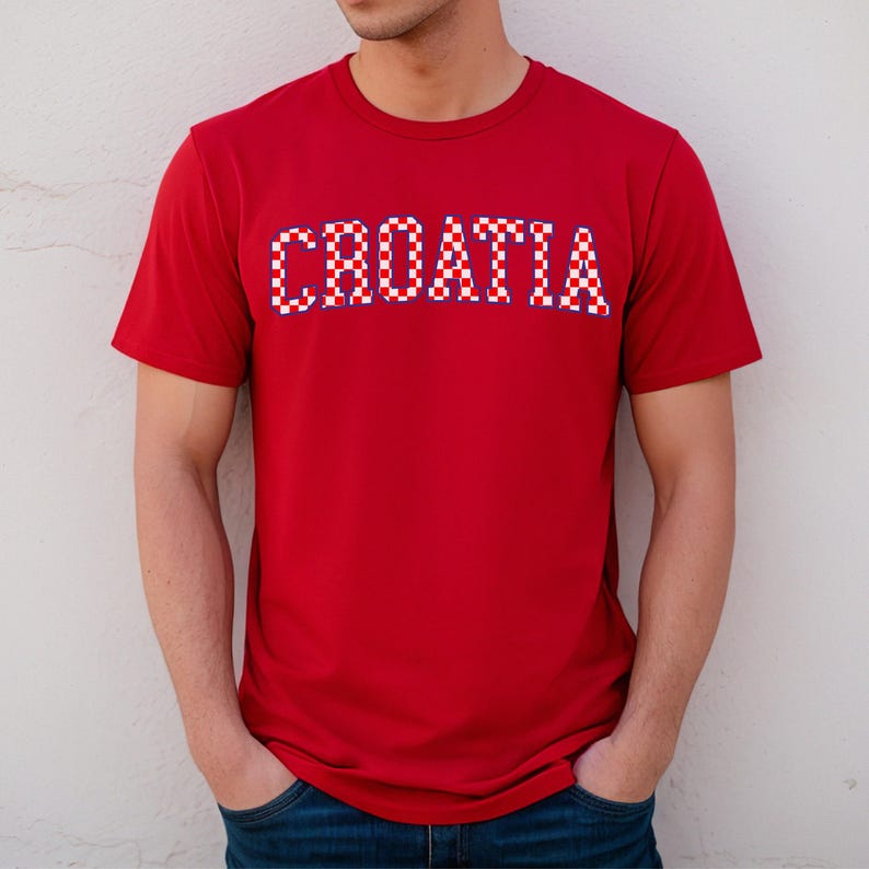 May include: A red t-shirt with the word "CROATIA" across the chest. The text is in a white, red, and blue checkered pattern, reminiscent of the Croatian flag. The shirt has short sleeves and a crew neck.