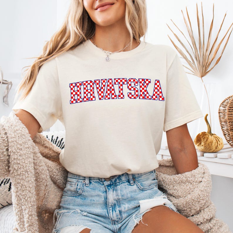 Pode incluir: Camiseta creme com a palavra "HRVATSKA" em letras xadrez vermelhas e brancas, contornadas em azul. A camiseta &eacute; de estilo casual, adequada para o uso di&aacute;rio.