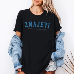 Op de afbeelding: Zwart T-shirt met een blauwe afbeelding met de tekst "ZMAJEVI" en daaronder in kleinere letters "BOSNA I HERCEGOVINA".