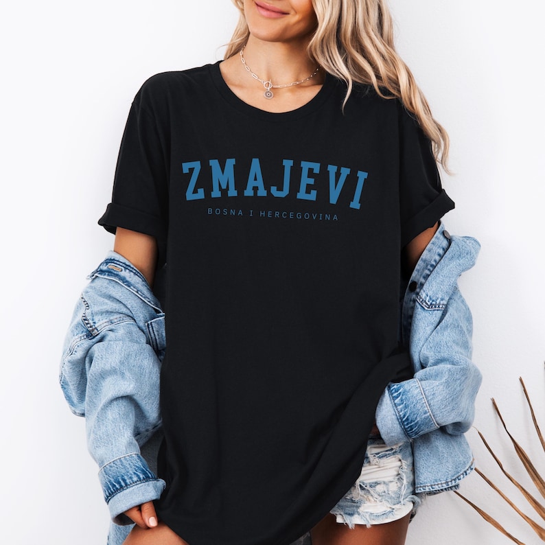 Op de afbeelding: Zwart T-shirt met een blauwe afbeelding met de tekst "ZMAJEVI" en daaronder in kleinere letters "BOSNA I HERCEGOVINA".