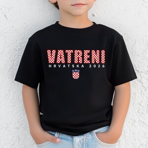 May include: Black t-shirt with the word "VATRENI" in red and white checkered letters. Below the word are the words "HRVATSKA 2026" and a Croatian coat of arms. The shirt is worn by a child.