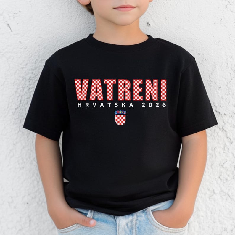 May include: Black t-shirt with the word "VATRENI" in red and white checkered letters. Below the word are the words "HRVATSKA 2026" and a Croatian coat of arms. The shirt is worn by a child.