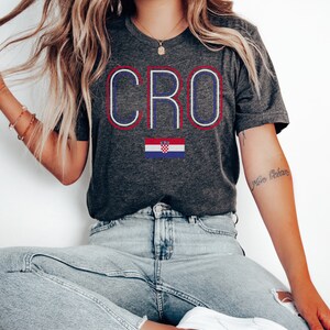 May include: A dark gray t-shirt with the word "CRO" in white, blue, and red outlined letters. A Croatian flag is below the word. The t-shirt is being worn by a person with long blonde hair and blue jeans.