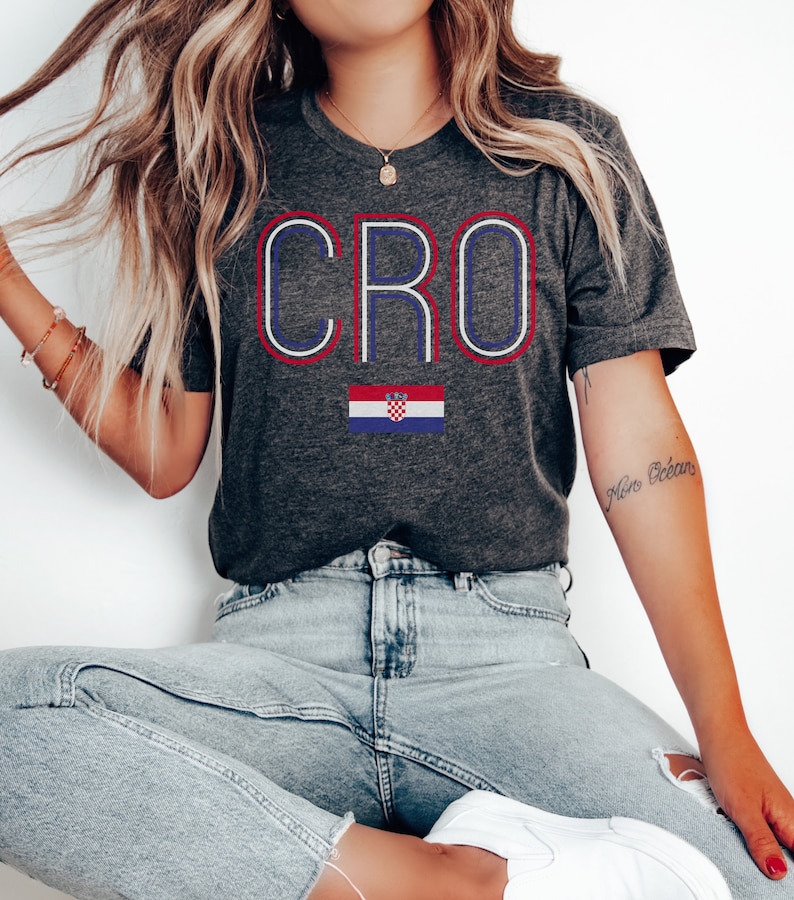 May include: A dark gray t-shirt with the word "CRO" in white, blue, and red outlined letters. A Croatian flag is below the word. The t-shirt is being worn by a person with long blonde hair and blue jeans.