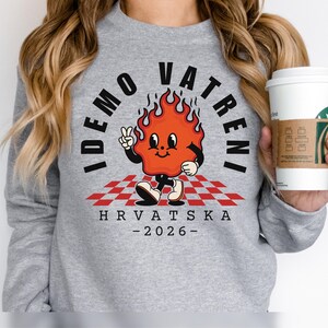 May include: A gray sweatshirt featuring a cartoon flame character with the text "IDEMO VATRENI" in a circular design. The character is on a red and white checkered pattern, with "HRVATSKA - 2026-" below.