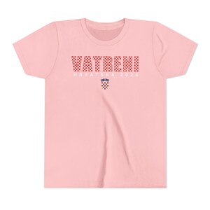 May include: A light pink t-shirt with the word "VATRENI" in a red and white checkered pattern across the chest. Below the word is the text "HRVATSKA 2026" and a Croatian coat of arms.
