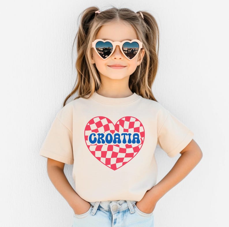 May include: A young person wearing a light beige t-shirt with a red and white checkered heart design and the word "CROATIA" in blue letters. The person is wearing heart-shaped sunglasses.