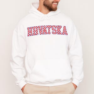 May include: White hooded sweatshirt with the word "HRVATSKA" in red and white checkered letters. The sweatshirt has a front pocket and a drawstring hood. The design is a nod to Croatian culture.