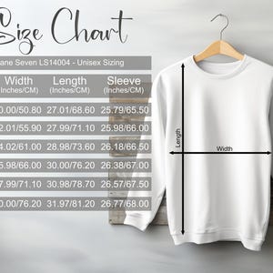 May include: White unisex sweatshirt hanging on a wooden hanger, with a size chart in the background. The size chart shows measurements in inches and centimeters for width, length, and sleeve length.