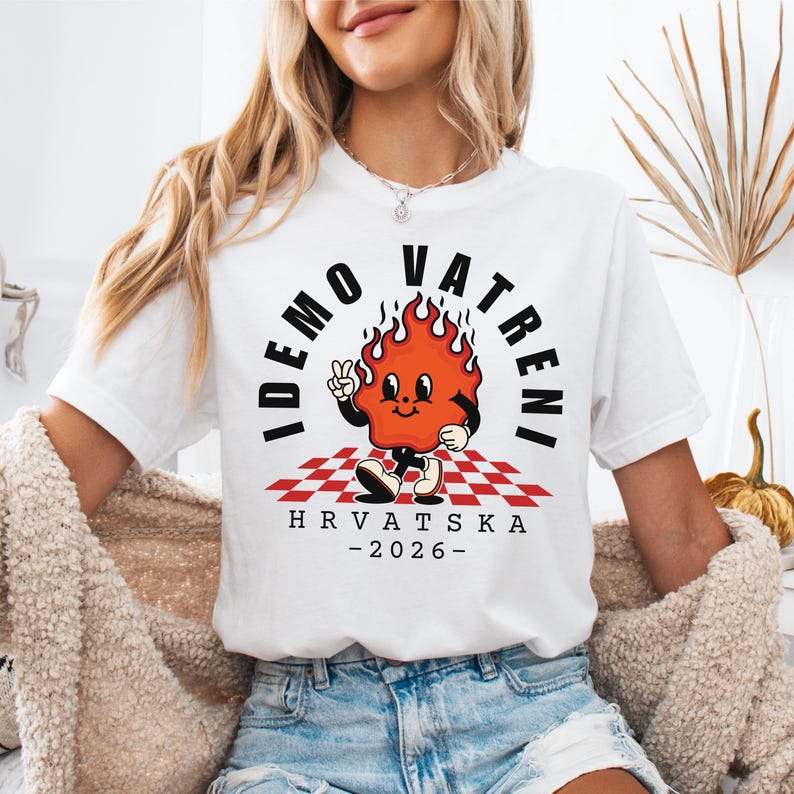 Puede incluir: Camiseta blanca con un personaje de llama de dibujos animados, el texto "IDEMO VATRENI" y "HRVATSKA -2026-". La llama es naranja y roja, de pie sobre un patr&oacute;n de cuadros rojos y blancos. Una camiseta casual.