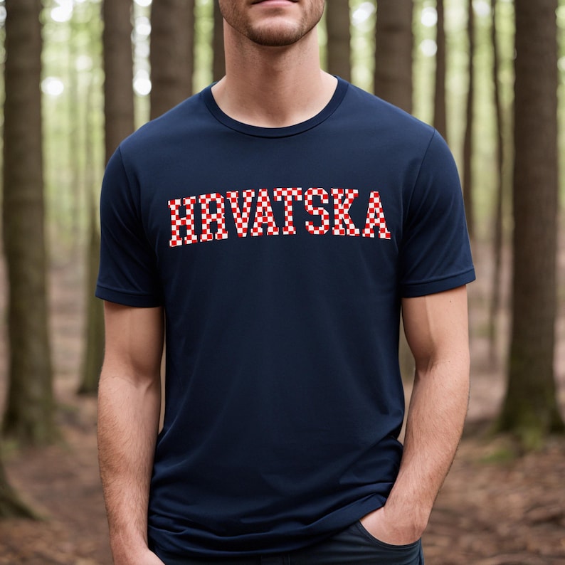 May include: A navy blue t-shirt featuring the word "HRVATSKA" in a red and white chequered design. The shirt has a crew neck and short sleeves. The background is a blurred forest scene.