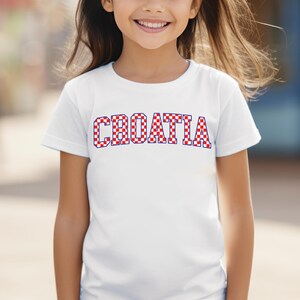 May include: A white t-shirt with the word "CROATIA" in red and white checkered letters. The shirt is short-sleeved and appears to be worn by a child. The background is blurred, suggesting an outdoor setting.