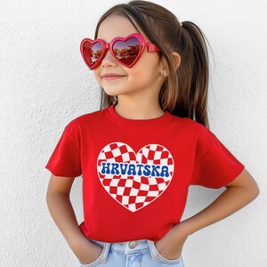 Puede incluir: Camiseta roja con un dise&ntilde;o de coraz&oacute;n a cuadros y la palabra "HRVATSKA" en letras azules. La camiseta la lleva puesta una ni&ntilde;a con gafas de sol rojas en forma de coraz&oacute;n.