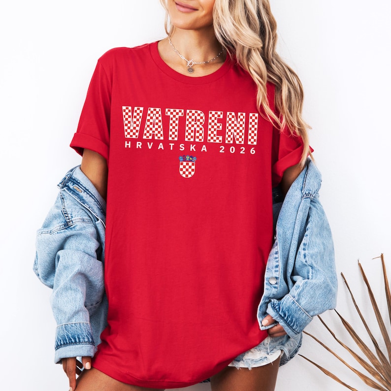 May include: A red t-shirt with the word "VATRENI" in white checkered letters. Below the word is "HRVATSKA 2026" and a Croatian coat of arms. The shirt is made of a soft material.