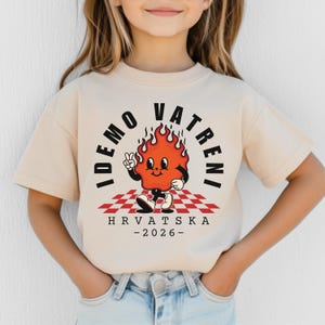 May include: A beige t-shirt featuring a cartoon flame character with the text "IDEMO VATRENI" and "HRVATSKA -2026-". The flame is on a red and white checkered pattern. The character is giving a peace sign.