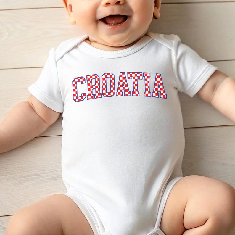 May include: A white baby bodysuit with the word "CROATIA" printed across the front in red and white checkered letters with a blue outline. The baby is smiling.