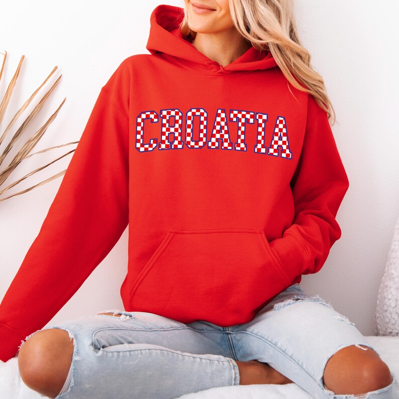 May include: A red hoodie with the word "CROATIA" in white letters with a checkered pattern. The hoodie has a front pocket and a hood. The person is wearing light blue jeans.