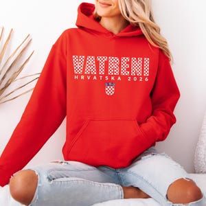 May include: A red hooded sweatshirt with the word "VATRENI" in white checkered letters. Below the word is "HRVATSKA 2026" and a Croatian coat of arms. The sweatshirt has a front pocket and a drawstring hood.