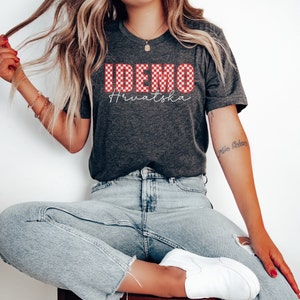May include: A gray t-shirt with a red and white checkered design that says "IDEMO Hrvatska". The shirt is being worn by a person with long blonde hair and blue jeans.