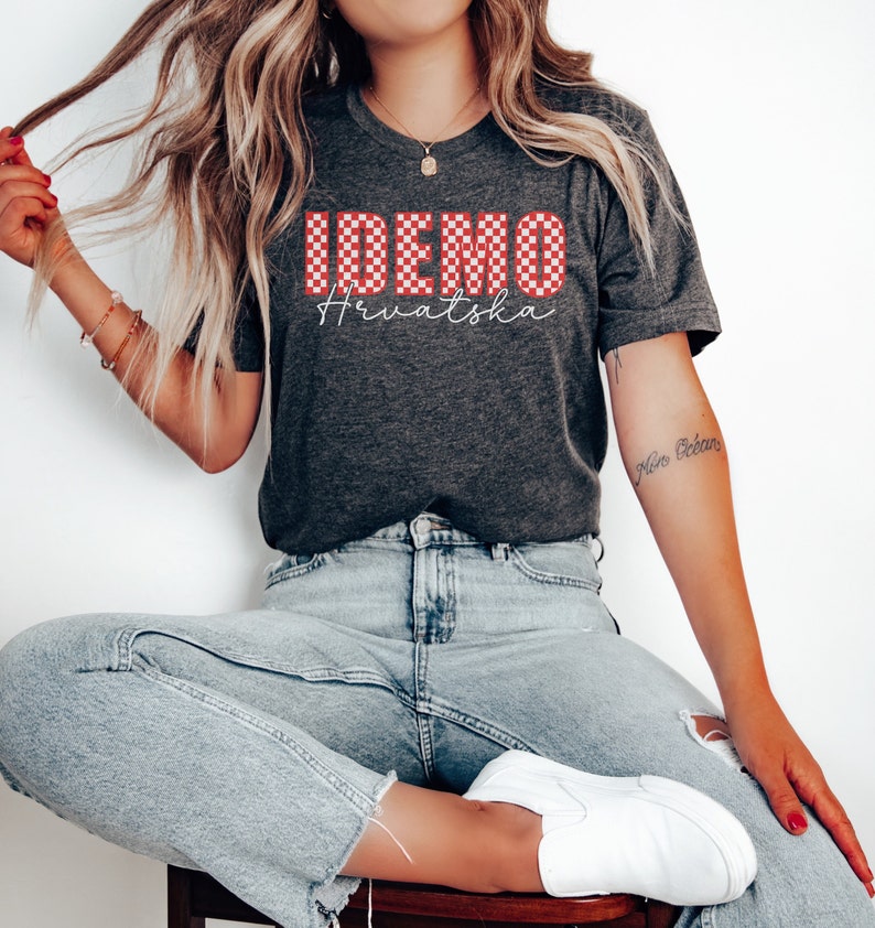 May include: A gray t-shirt with a red and white checkered design that says "IDEMO Hrvatska". The shirt is being worn by a person with long blonde hair and blue jeans.