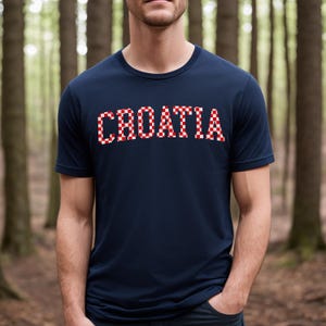 May include: Navy blue t-shirt with the word "CROATIA" in red and white checkered letters across the chest. The shirt has short sleeves and a crew neck. The background is a blurred forest scene.