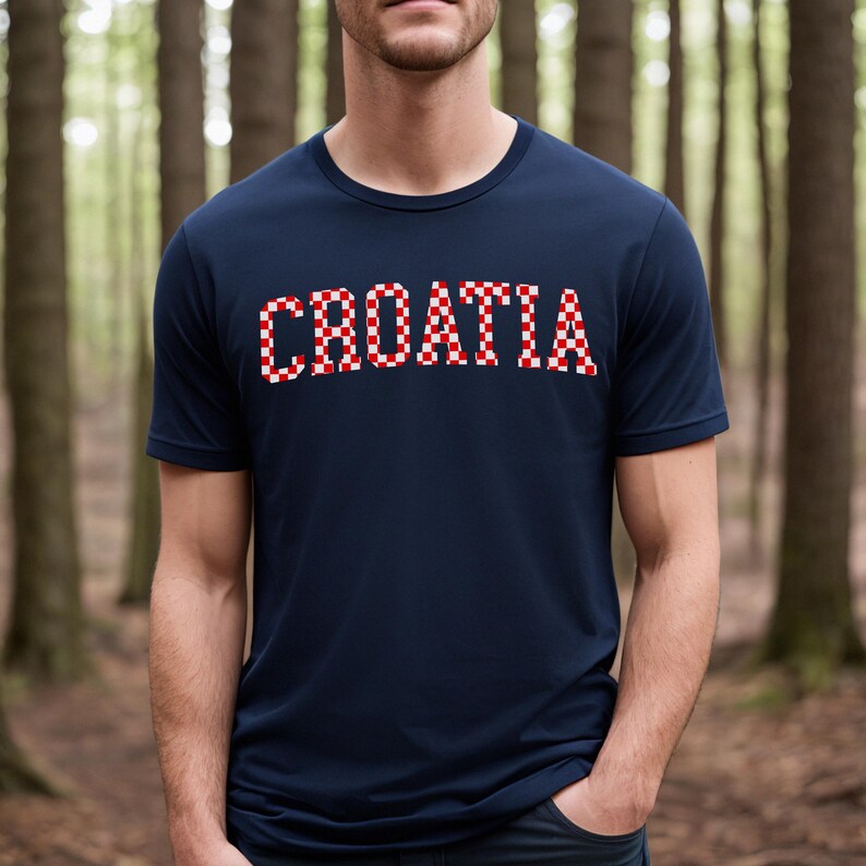May include: Navy blue t-shirt with the word "CROATIA" in red and white checkered letters across the chest. The shirt has short sleeves and a crew neck. The background is a blurred forest scene.