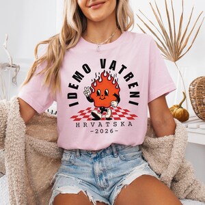 Puede incluir: Camiseta rosa claro con un personaje de llama de dibujos animados y el texto "IDEMO VATRENI" y "HRVATSKA -2026-". El personaje de la llama est&aacute; sobre un patr&oacute;n de cuadros rojos y blancos.