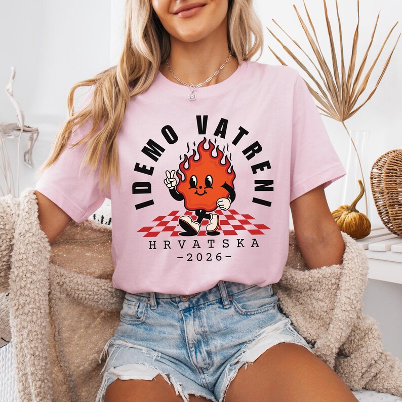 Puede incluir: Camiseta rosa claro con un personaje de llama de dibujos animados y el texto "IDEMO VATRENI" y "HRVATSKA -2026-". El personaje de la llama est&aacute; sobre un patr&oacute;n de cuadros rojos y blancos.