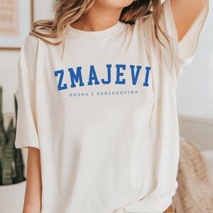 Op de afbeelding: Een wit T-shirt met een blauwe afbeelding waarop "ZMAJEVI" en "BOSNA I HERCEGOVINA" in een college-lettertype staan.