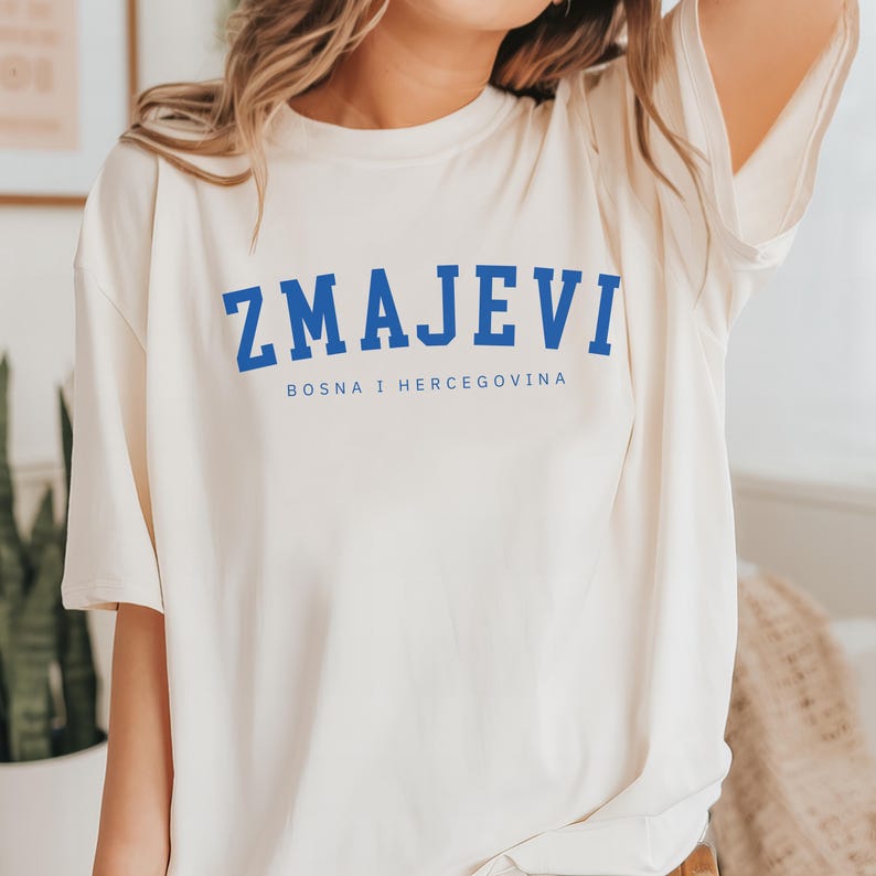 Op de afbeelding: Een wit T-shirt met een blauwe afbeelding waarop "ZMAJEVI" en "BOSNA I HERCEGOVINA" in een college-lettertype staan.